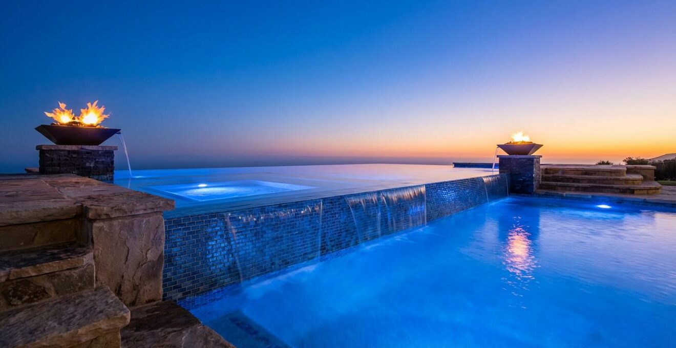Twilight view of luxury pool with infinity edge spa, fire bowls, and illuminated water features